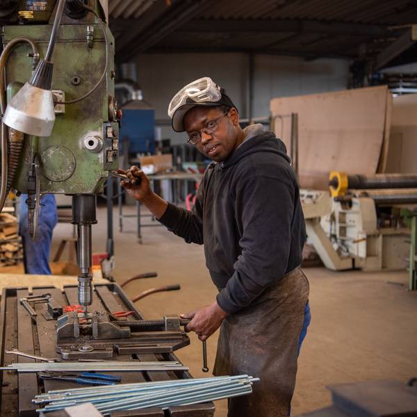 Hady Bah steht an einem Gerät in einer Metallwerkstatt. Er trägt eine Lederschürze und eine Sicherheitsbrille.