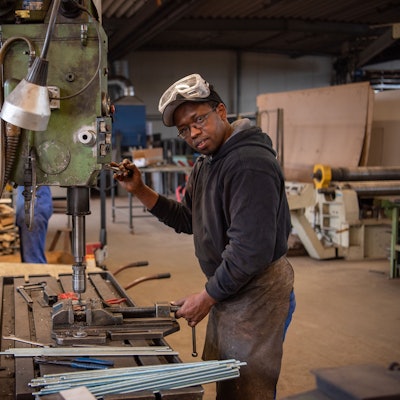 Hady Bah steht an einem Gerät in einer Metallwerkstatt. Er trägt eine Lederschürze und eine Sicherheitsbrille.