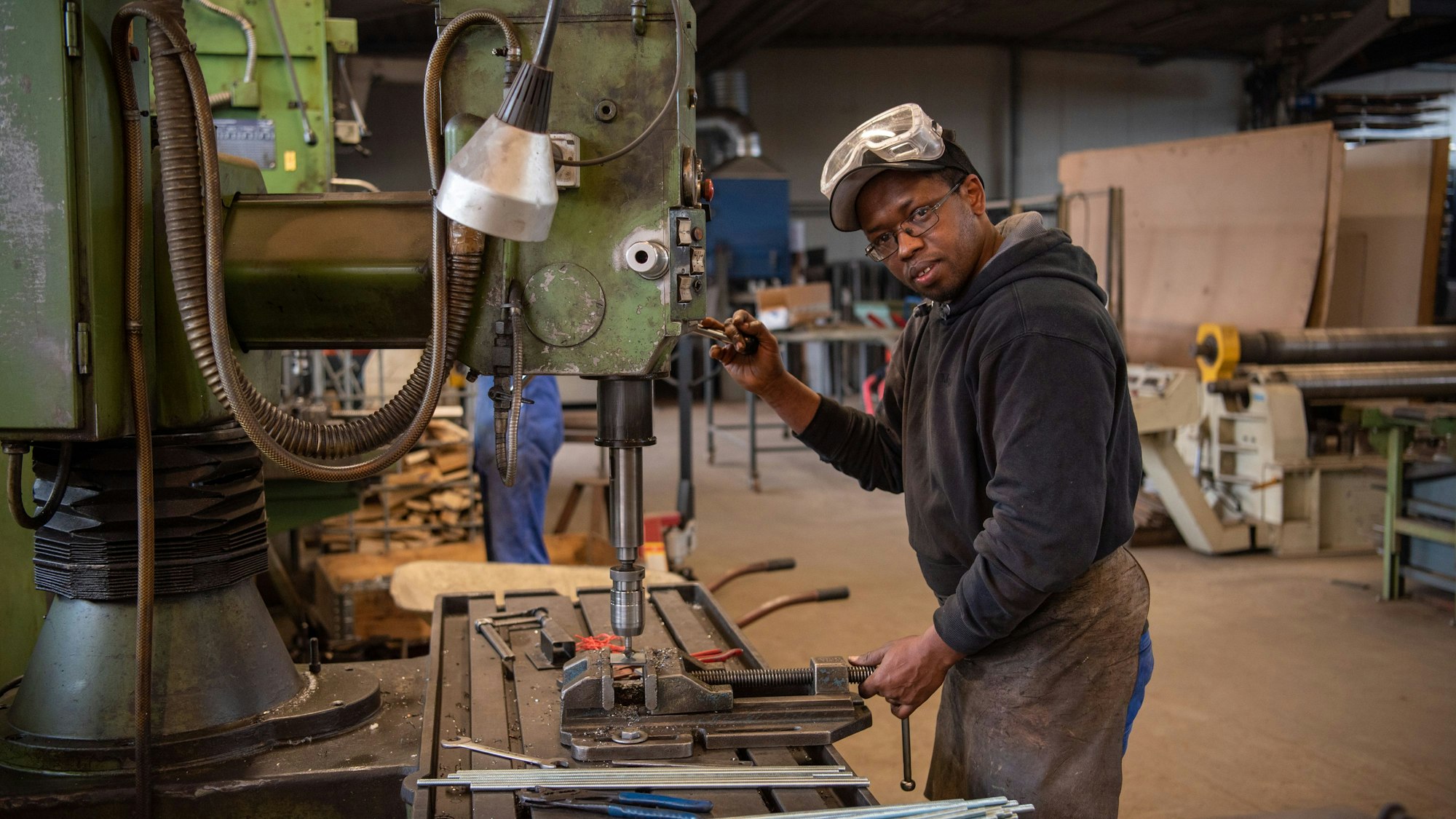 Hady Bah steht an einem Gerät in einer Metallwerkstatt. Er trägt eine Lederschürze und eine Sicherheitsbrille.