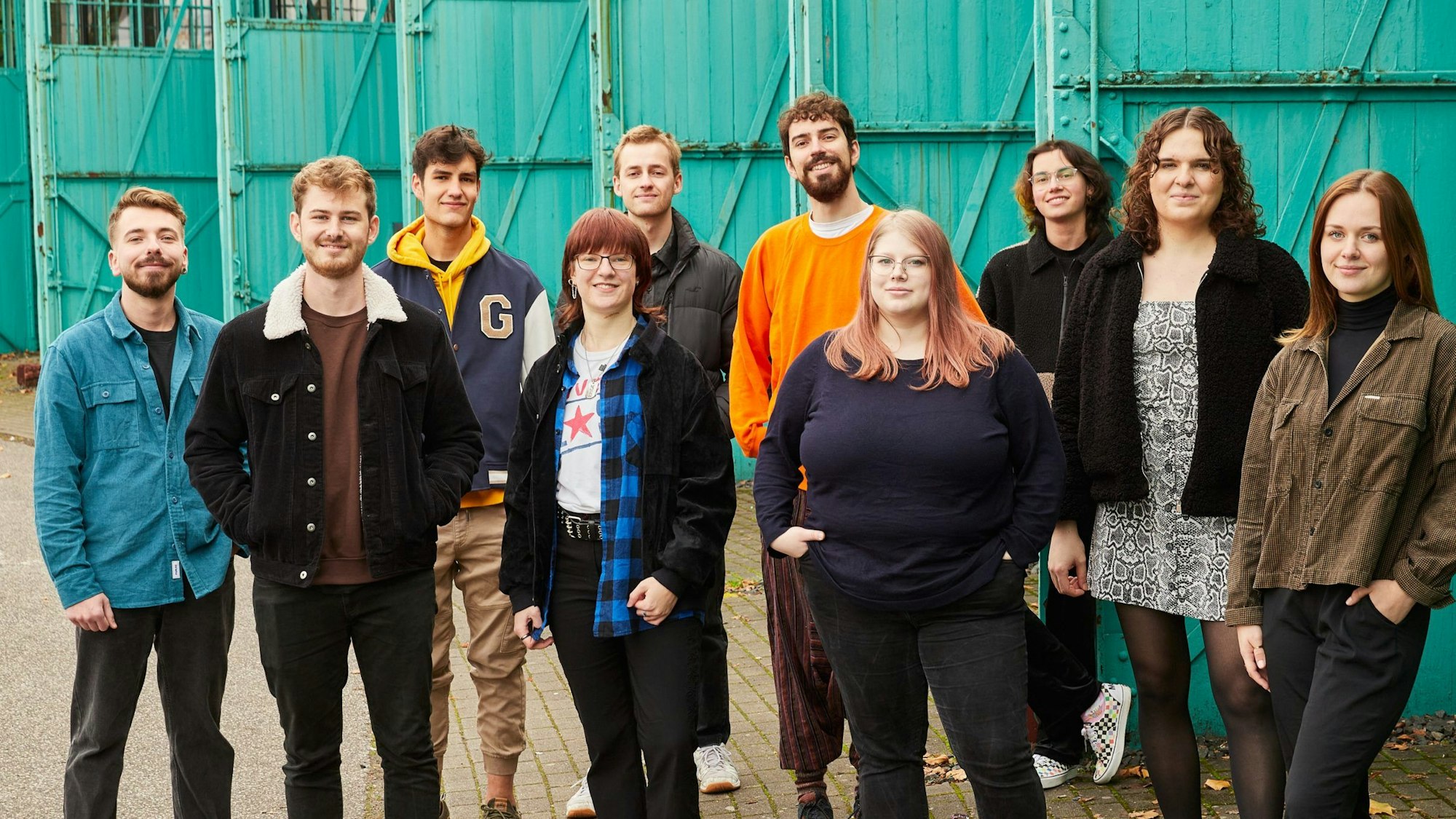 Zehn junge queere Beraterinnen und Berater des neuen Kölner Messenger-Tools „Coming out und..." stehen vor grünen Toren einer Industriehalle, sie beraten online Jugendliche in ähnlichen Lebenssituationen oder-lagen.