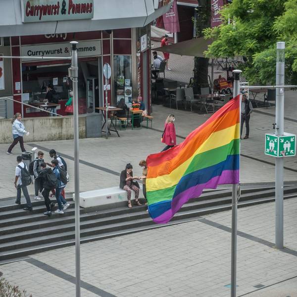 Im Juni 2021 weht auf dem Rathausvorplatz in Wiesdorf eine Regenbogenfahne. Passanten flanieren, Schüler stehen beieinander.