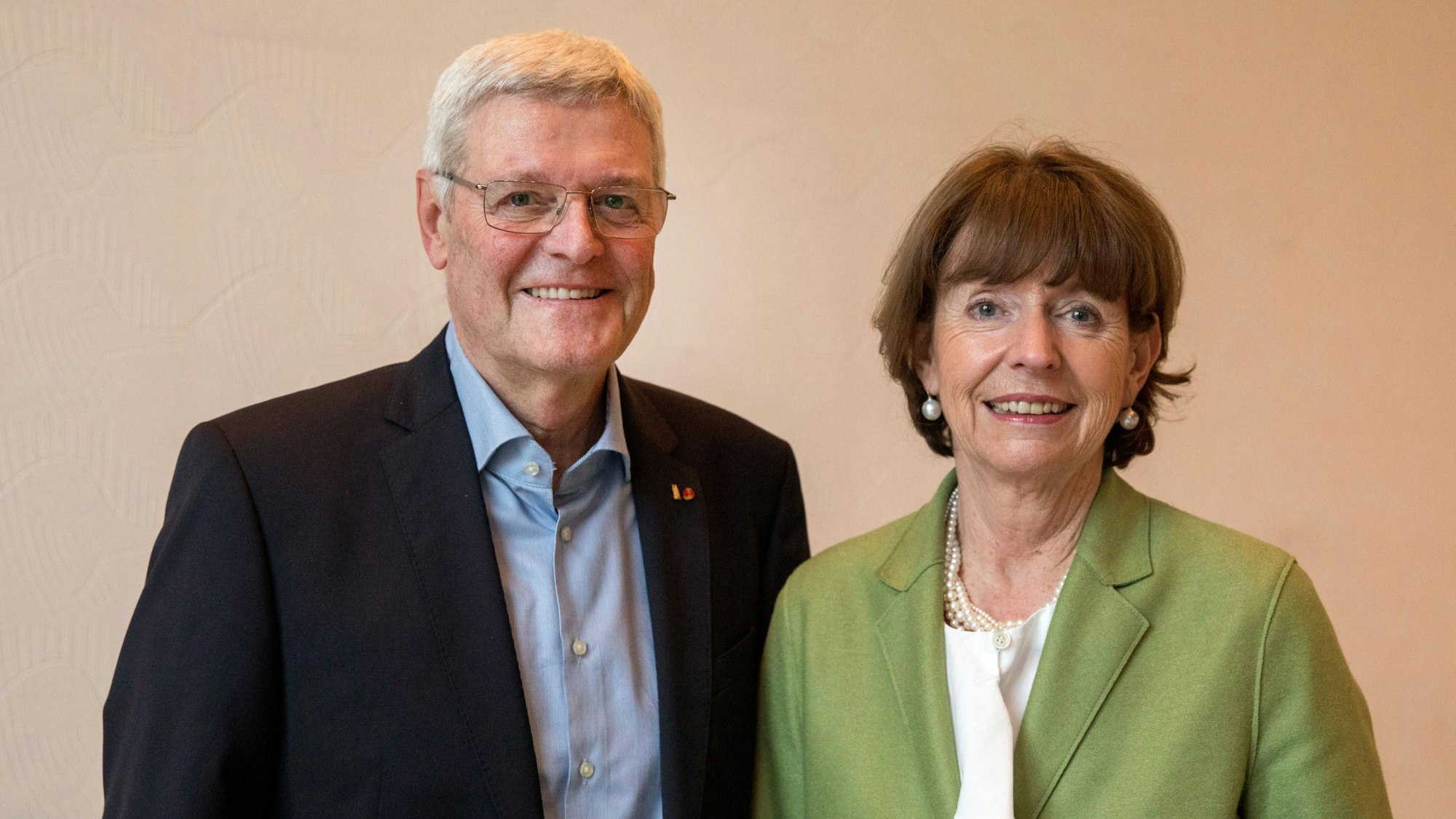 Die beiden blicken in die Kamera vor einem hellen Hintergrund.