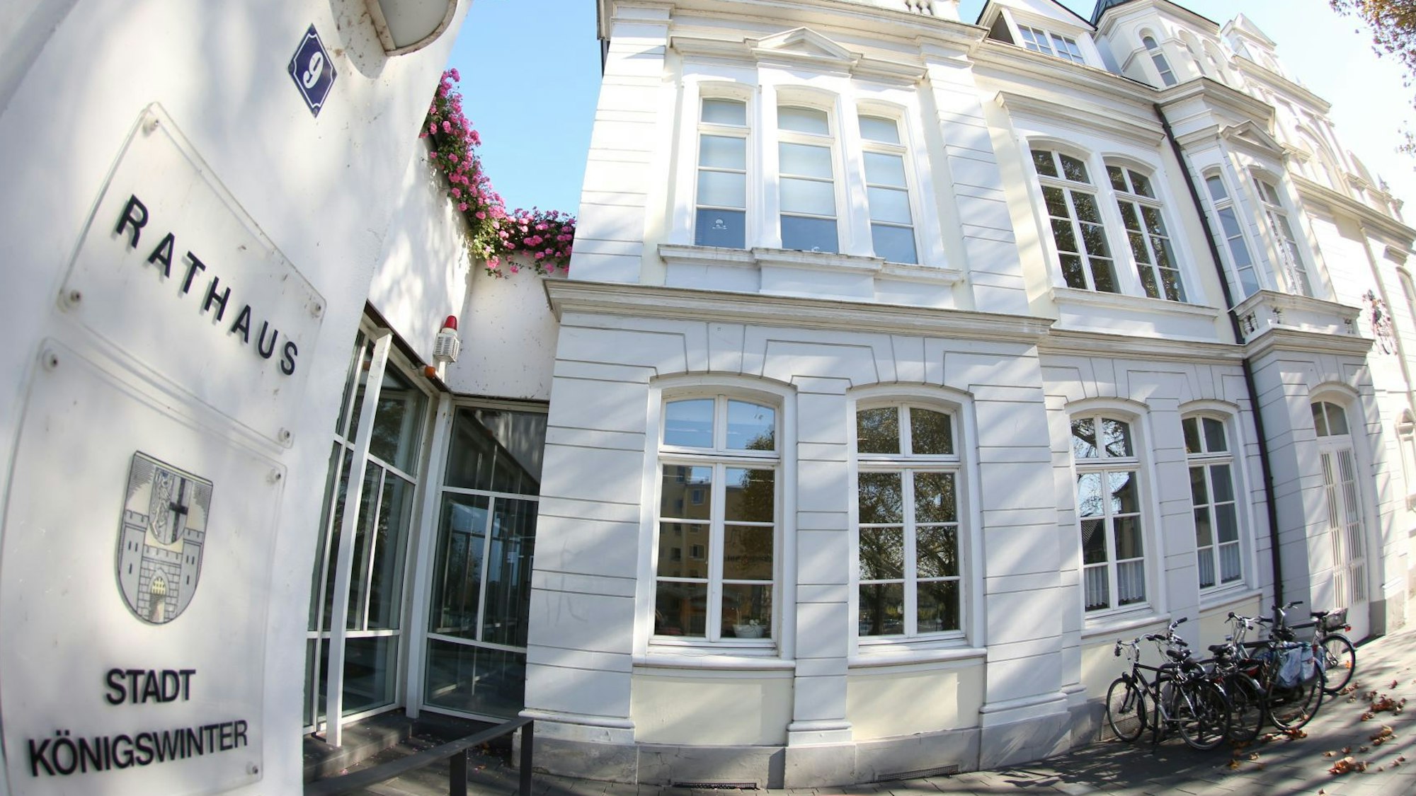 Ein Schild mit dem Stadtwappen und den Schriftzügen „Rathaus Stadt Königswinter“ hängt links am Gebäude. Vor dem Rathaus sind einige Fahrräder abgestellt worden.