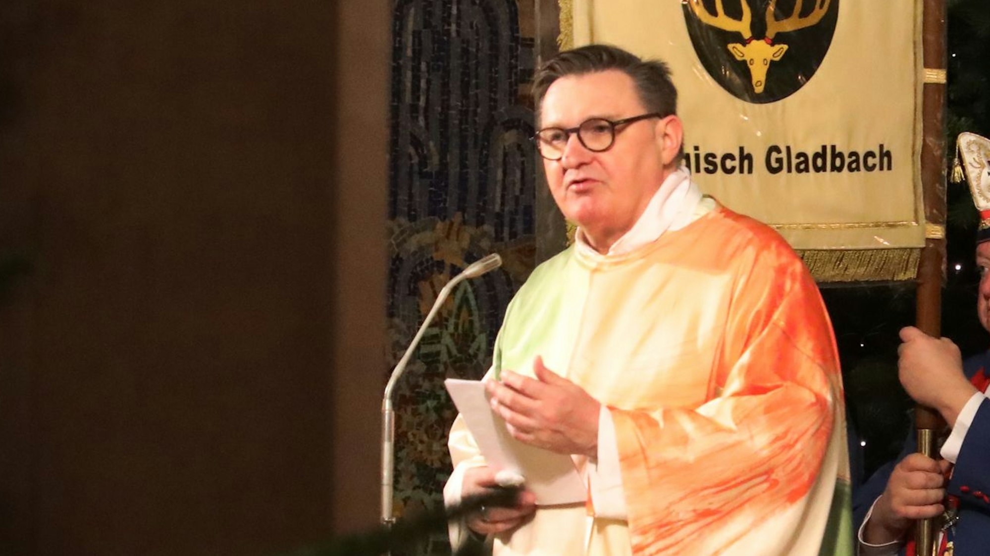 Kreisdechant Norbert Hörter steht im Messgewand vor einer Fahne, auf der das Bergisch Gladbacher Stadtwappen und der Schriftzug des Kreisstadtnamens zu sehen sind.