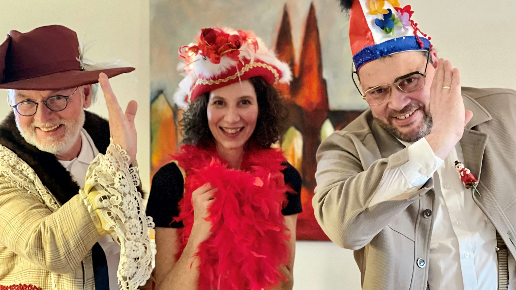 Ein Mann mit weißem Bart und Brille trägt Schlapphut und ein helle, karierte Jacke mit Rüschenärmel, eine dunkelhaarige Frau eine rote Federboa und einen roten Dreispitz, ein weiterer Mann eine blau-rote Narrenkappe. Sie heben die Hand an die Stirn zum jecken Gruß,
