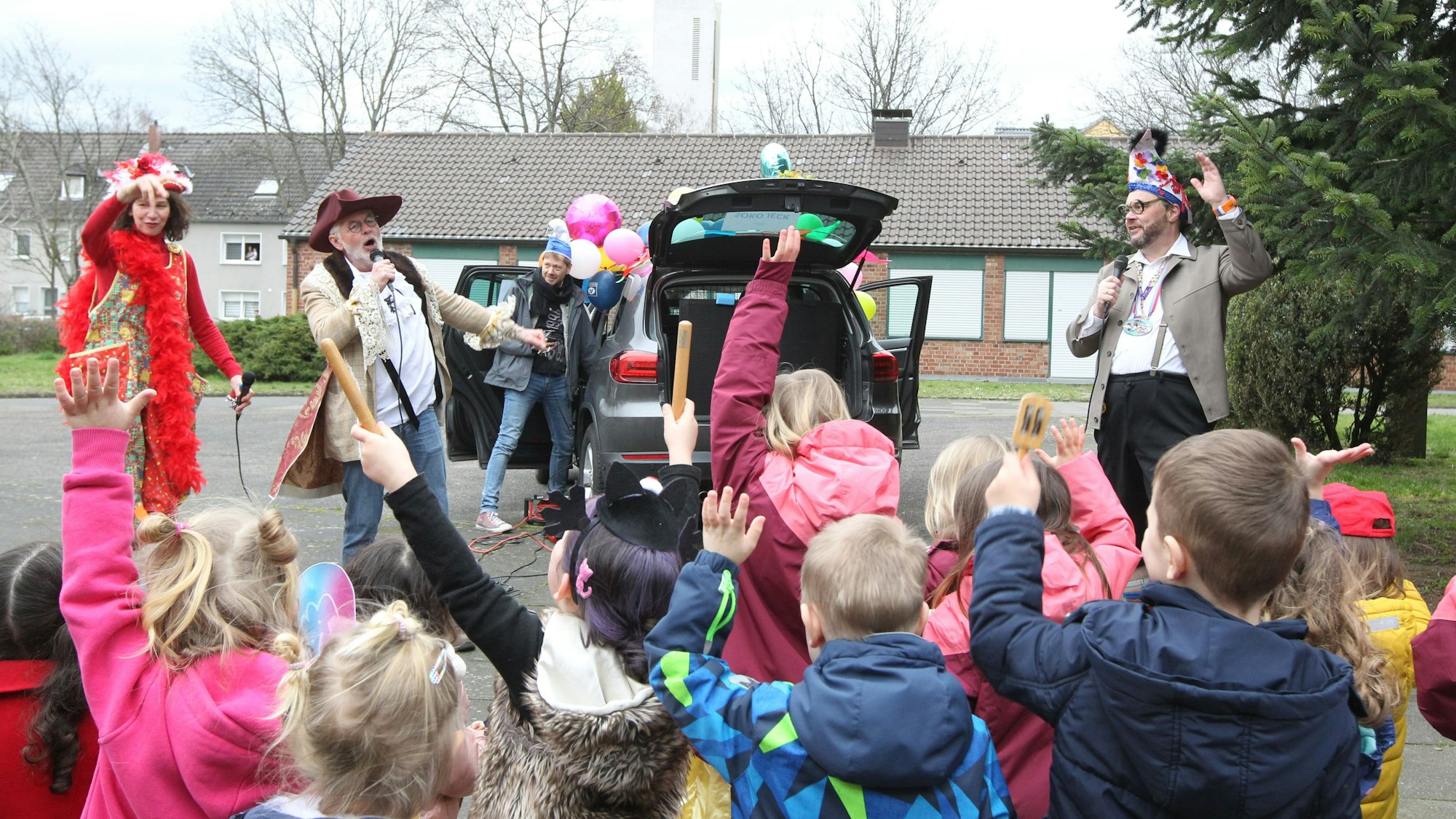 Kinder, zum Teil in Kostümen, reißen die Arme hoch, während vor ihnen kostümierten Männer und eine Frau an einem mit Luftballons geschmückten Auto in Mikrofone singen.