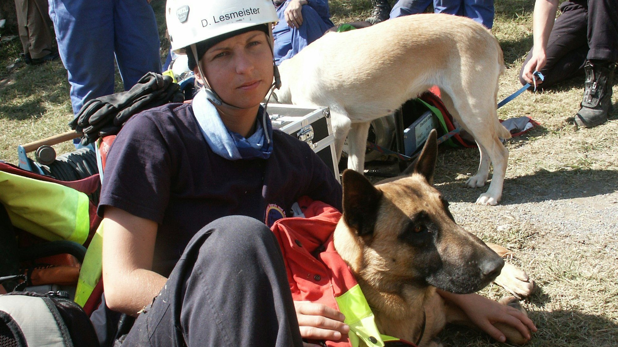 Daniela Lesmeister sitzt auf trockenem Gras, trägt einen weißen Schutzhelm, T-Shirt und weite Hose. Ihren Arm hat sie um den Schäferhund Amoc gelegt, der neben ihr liegt.