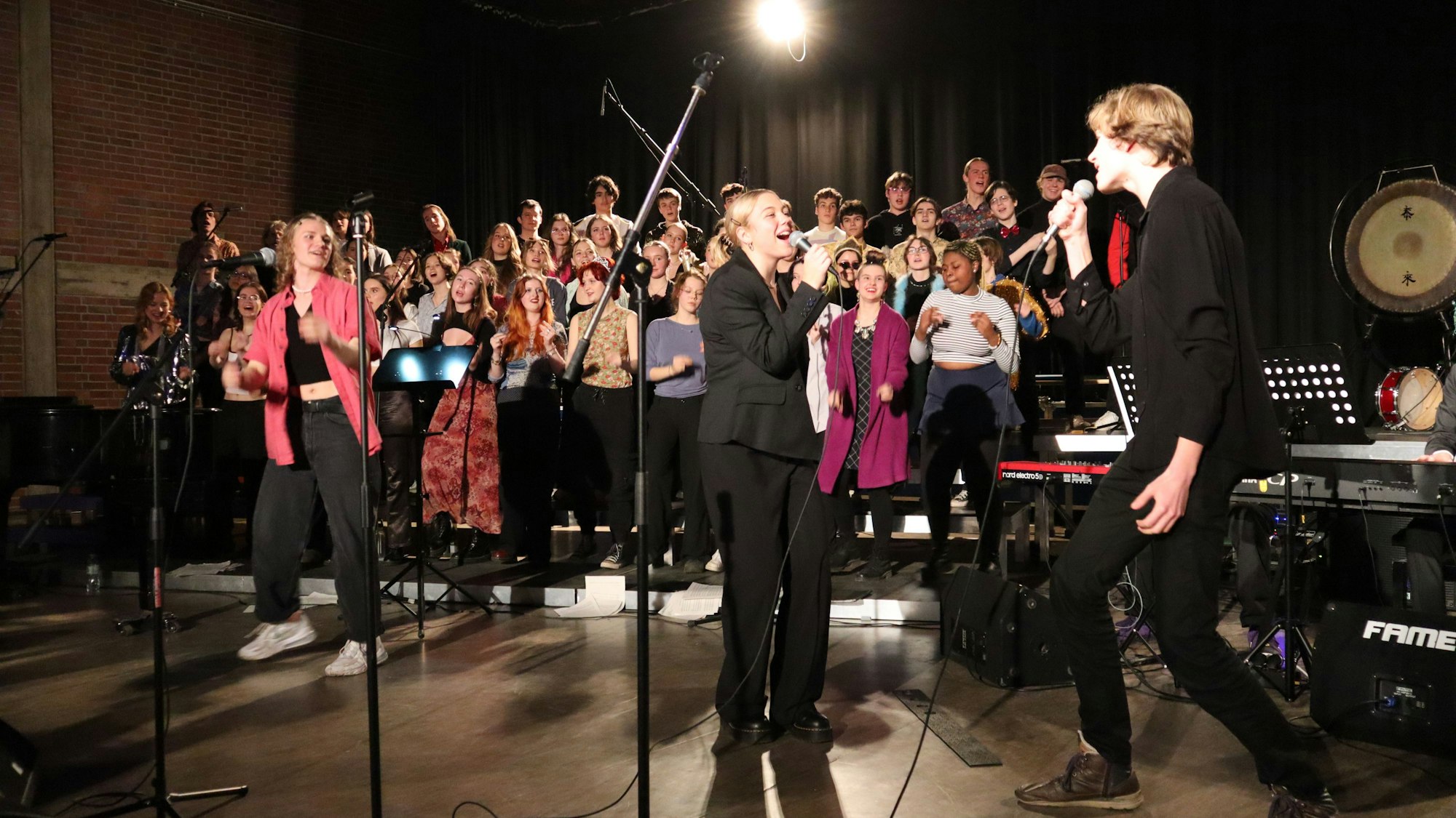 Ein Chor und drei einzelne Sänger stehen auf einer Bühne, ein Scheinwerfer leuchtet in der Mitte.