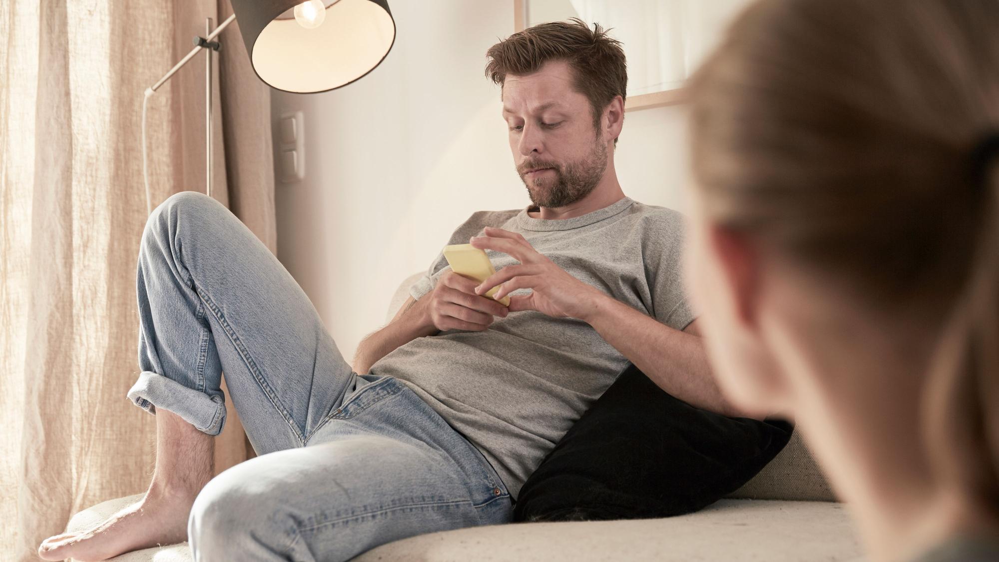 Eine Frau schaut einen Mann an, der mit etwas Abstand auf dem Sofa sitzt und etwas gelangweilt auf sein Handy blickt.