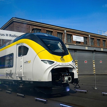 Ein Wasserstoffzug verlässt eine Werkshalle, zu sehen ist der erste Wagen, er ist weiß mit einem gelben Streifen und einem schwarzen Dach. Im Hintergrund ist eine historische Fabrik-Fassade zu sehen.