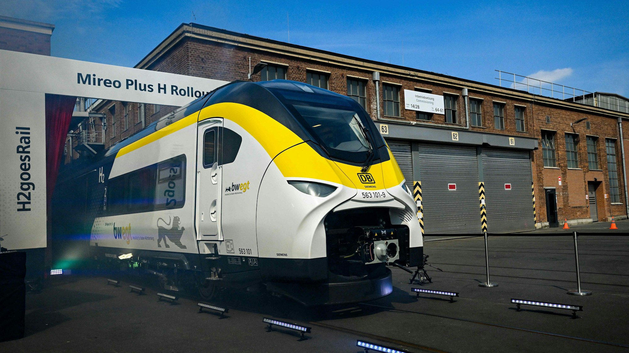 Ein Wasserstoffzug verlässt eine Werkshalle, zu sehen ist der erste Wagen, er ist weiß mit einem gelben Streifen und einem schwarzen Dach. Im Hintergrund ist eine historische Fabrik-Fassade zu sehen.