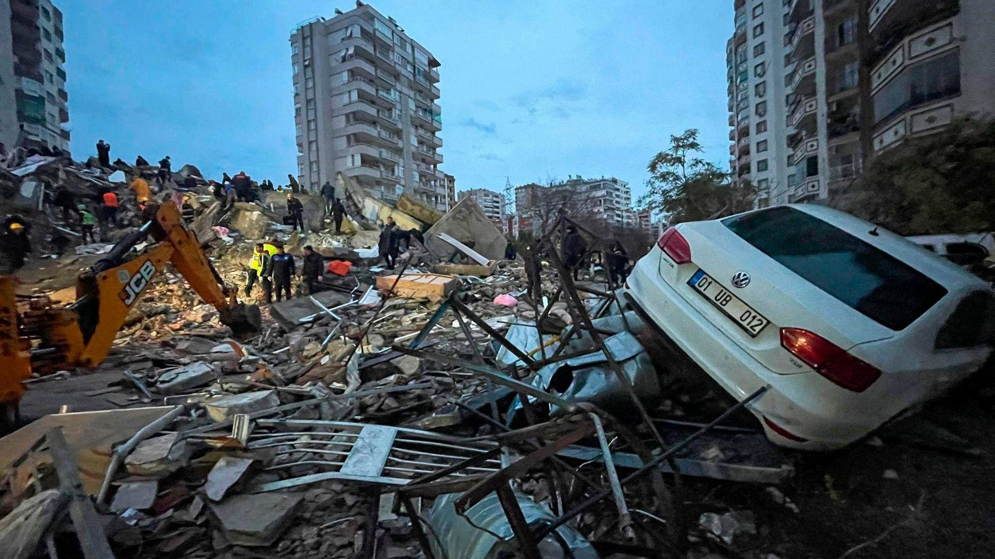 Notfallteams suchen in der türkischen Stadt Adana nach Menschen in einem zerstörten Gebäude, während im Bildvordergrund rechts ein Auto auf den Trümmern liegt.