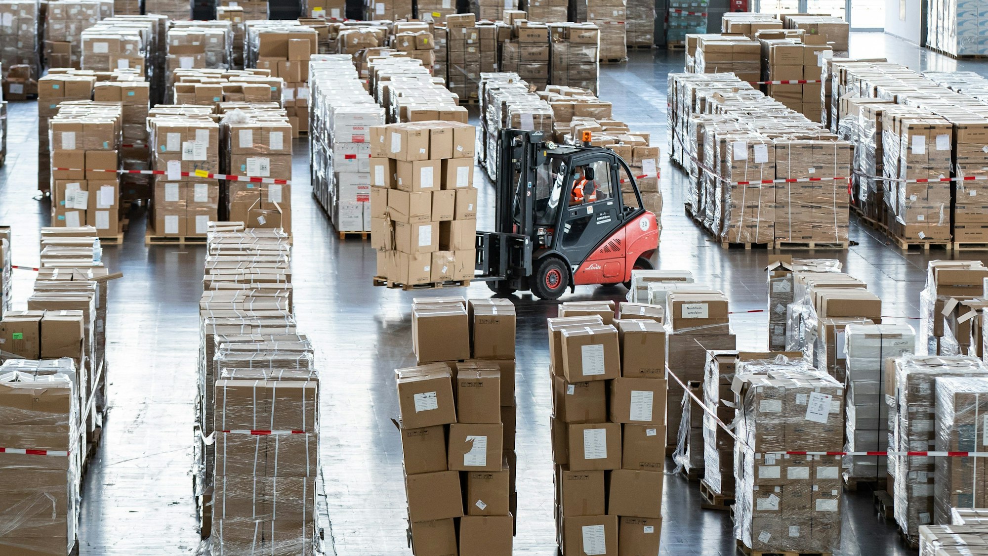 In einer Lagerhalle stapeln sich unzählige Kartons. Ein Gabelstapler mit Kartons voll Schutzausrüstung fährt durch die Halle.