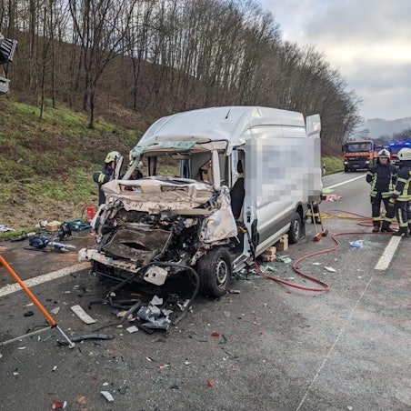 Ein schwer beschädigter Kastenwagen steht auf der Autobahn. Teile des Fahrzeugs liegen auf der Fahrbahn. Feuerwehrleute stehen neben dem Wagen.