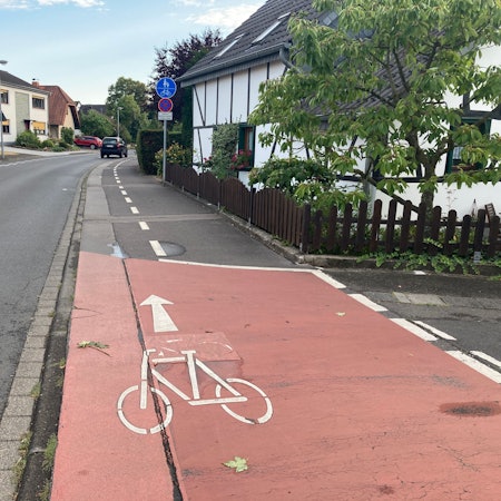 Bachstraße in Lohmar. Ein Fahrradweg ist rechts von der Straße zu sehen. Im Hintergrund stehen Häuser und Autos.
