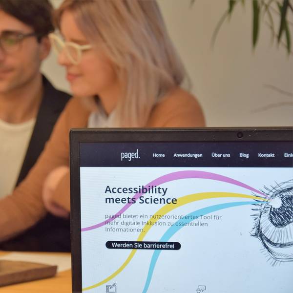 Deniz Tuzsus und Dana Pietralla sitzen mit Laptop, Notizheft und Handy an einem Tisch und sind verschwommen. Im Vordergrund ist ihre Website paged. auf einem weiteren Laptop zu sehen. Die Website hat einen weißen Hintergrund mit drei geschwungenen Linien in pink, gelb und hellblau, die von einem gezeichneten Auge ausgehen. „Accessibility meets Science“ und „Werden Sie barrierefrei“ stehen darauf geschrieben.