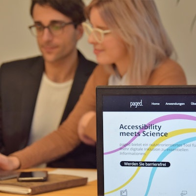 Deniz Tuzsus und Dana Pietralla sitzen mit Laptop, Notizheft und Handy an einem Tisch und sind verschwommen. Im Vordergrund ist ihre Website paged. auf einem weiteren Laptop zu sehen. Die Website hat einen weißen Hintergrund mit drei geschwungenen Linien in pink, gelb und hellblau, die von einem gezeichneten Auge ausgehen. „Accessibility meets Science“ und „Werden Sie barrierefrei“ stehen darauf geschrieben.