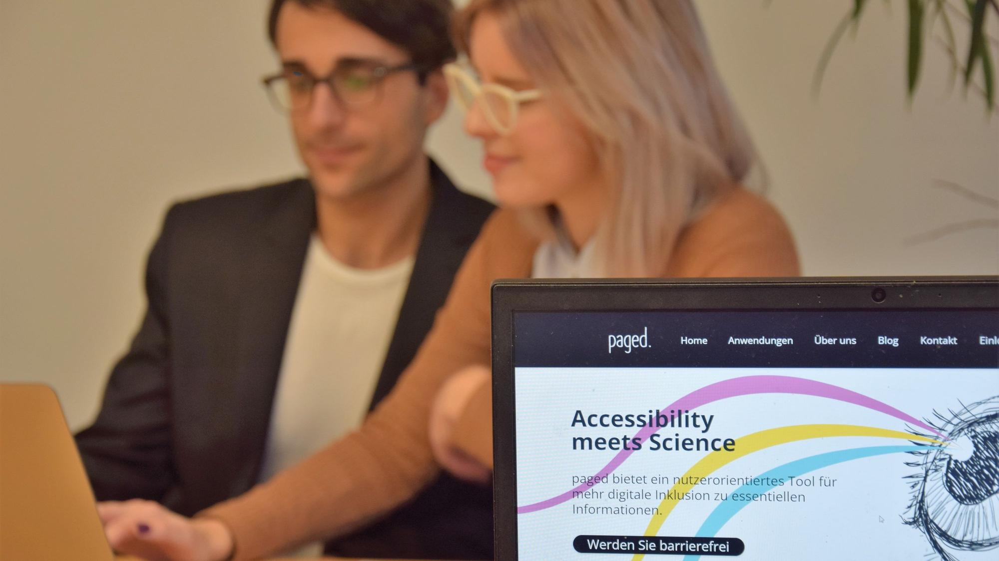 Deniz Tuzsus und Dana Pietralla sitzen mit Laptop, Notizheft und Handy an einem Tisch und sind verschwommen. Im Vordergrund ist ihre Website paged. auf einem weiteren Laptop zu sehen. Die Website hat einen weißen Hintergrund mit drei geschwungenen Linien in pink, gelb und hellblau, die von einem gezeichneten Auge ausgehen. „Accessibility meets Science“ und „Werden Sie barrierefrei“ stehen darauf geschrieben.