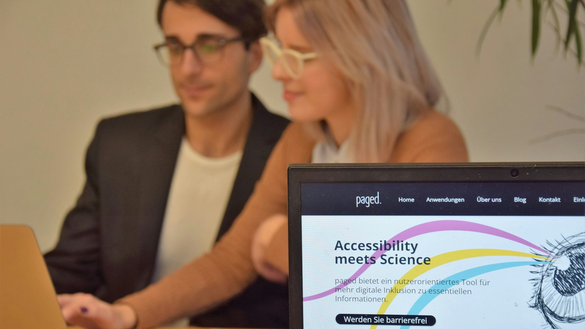 Deniz Tuzsus und Dana Pietralla sitzen mit Laptop, Notizheft und Handy an einem Tisch und sind verschwommen. Im Vordergrund ist ihre Website paged. auf einem weiteren Laptop zu sehen. Die Website hat einen weißen Hintergrund mit drei geschwungenen Linien in pink, gelb und hellblau, die von einem gezeichneten Auge ausgehen. „Accessibility meets Science“ und „Werden Sie barrierefrei“ stehen darauf geschrieben.