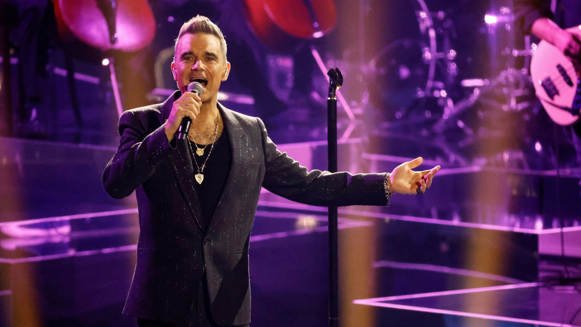 Robbie Williams steht in einem schwarzen Anzug auf der Bühne.
