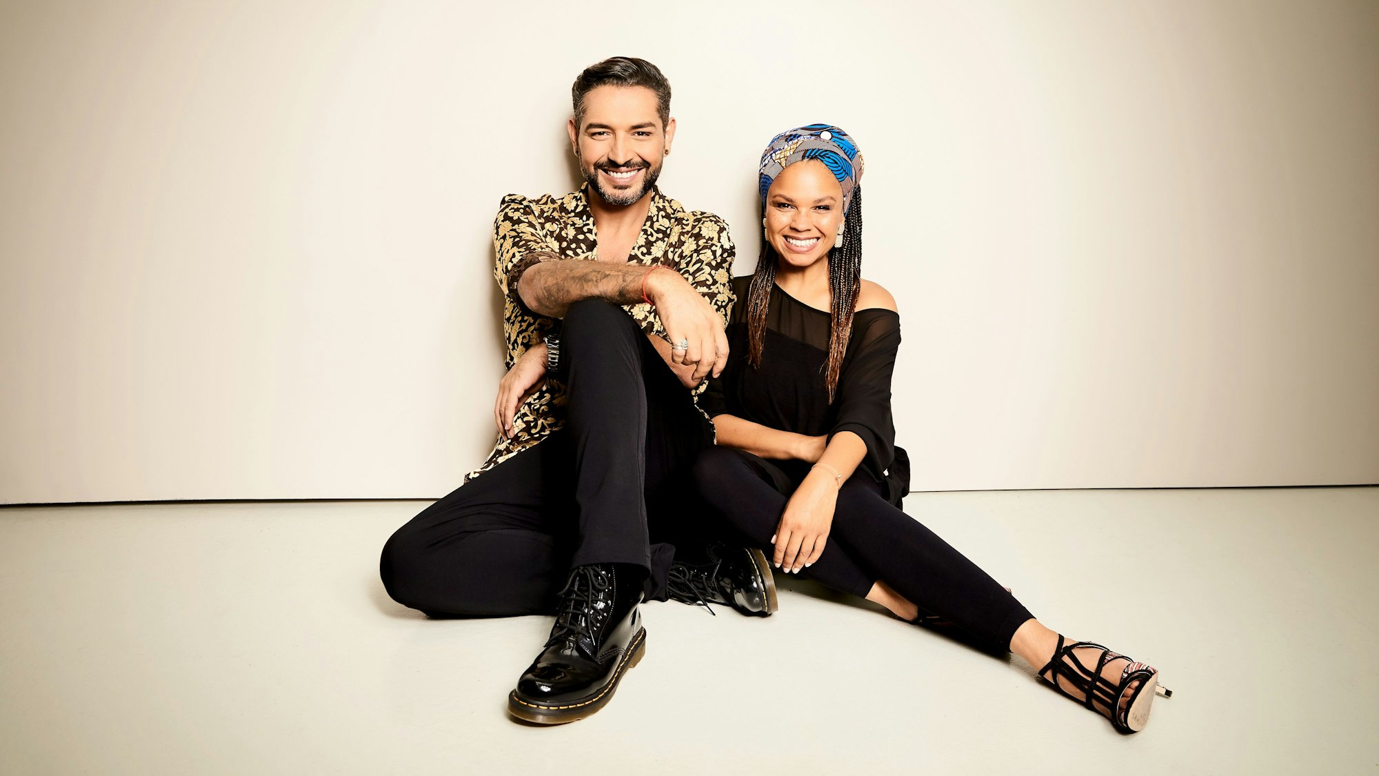 Gianni Jovanovic und Oyindamola Alashe sitzen nebeneinander auf dem Boden und lächeln in die Kamera.