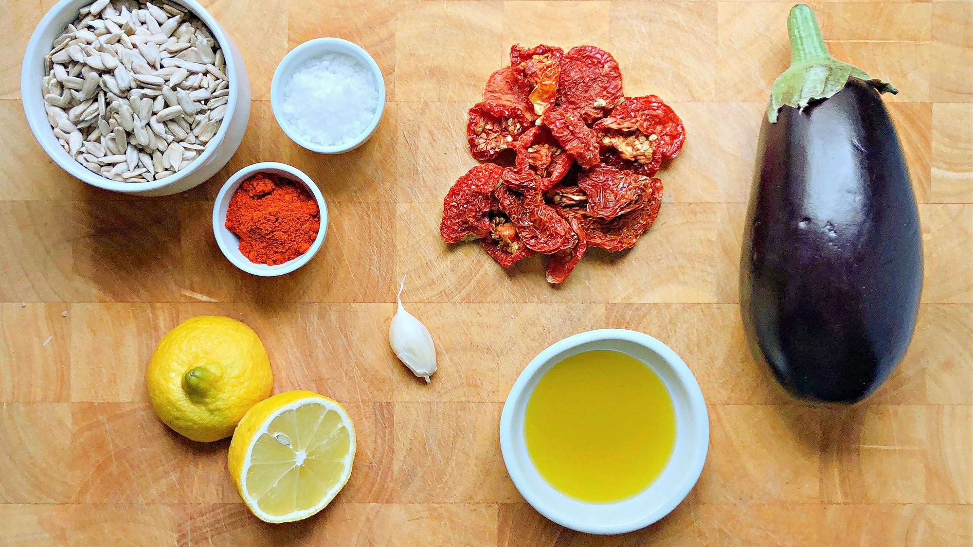 Zutaten für einen aufstrich liegen auf einer hölzernen Tischplatte, unter anderem: Sonnenblumenkerne, getrocknete Tomaten, Aubergine, Knoblauch, Paprikapulver, Salz und Öl sowie etwas Zitronensaft.