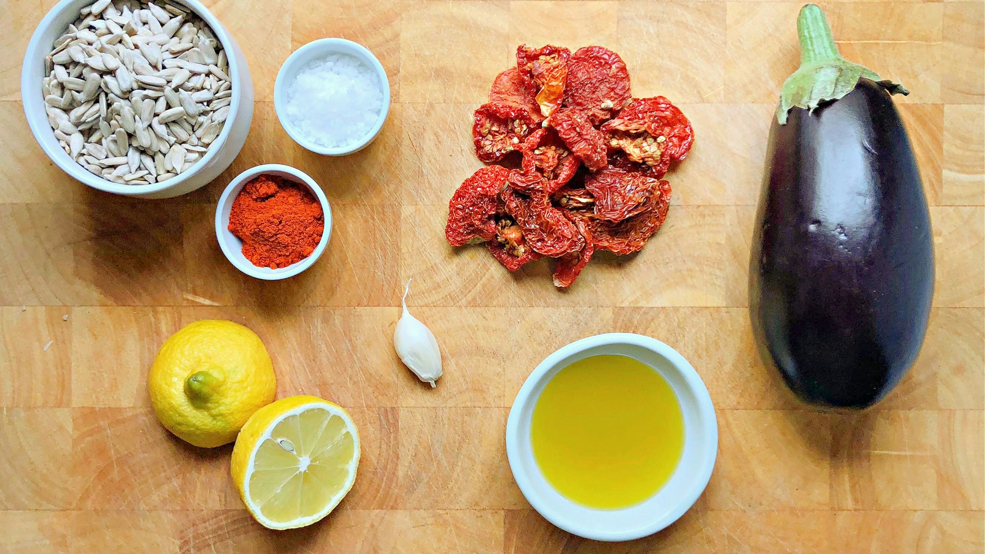 Zutaten für einen aufstrich liegen auf einer hölzernen Tischplatte, unter anderem: Sonnenblumenkerne, getrocknete Tomaten, Aubergine, Knoblauch, Paprikapulver, Salz und Öl sowie etwas Zitronensaft.