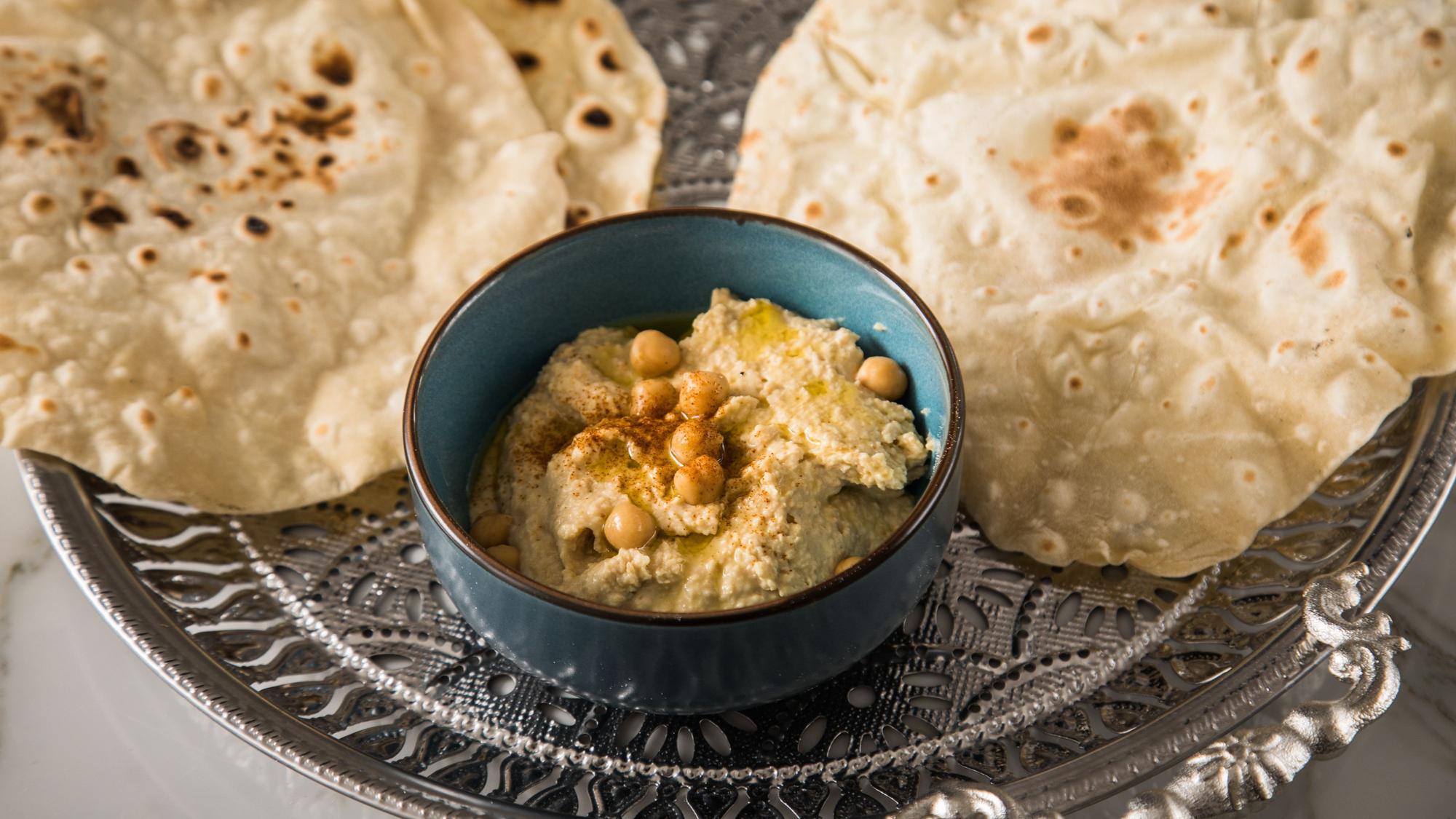 Hummus in einem Schälchen, daneben zwei Fladenbrote