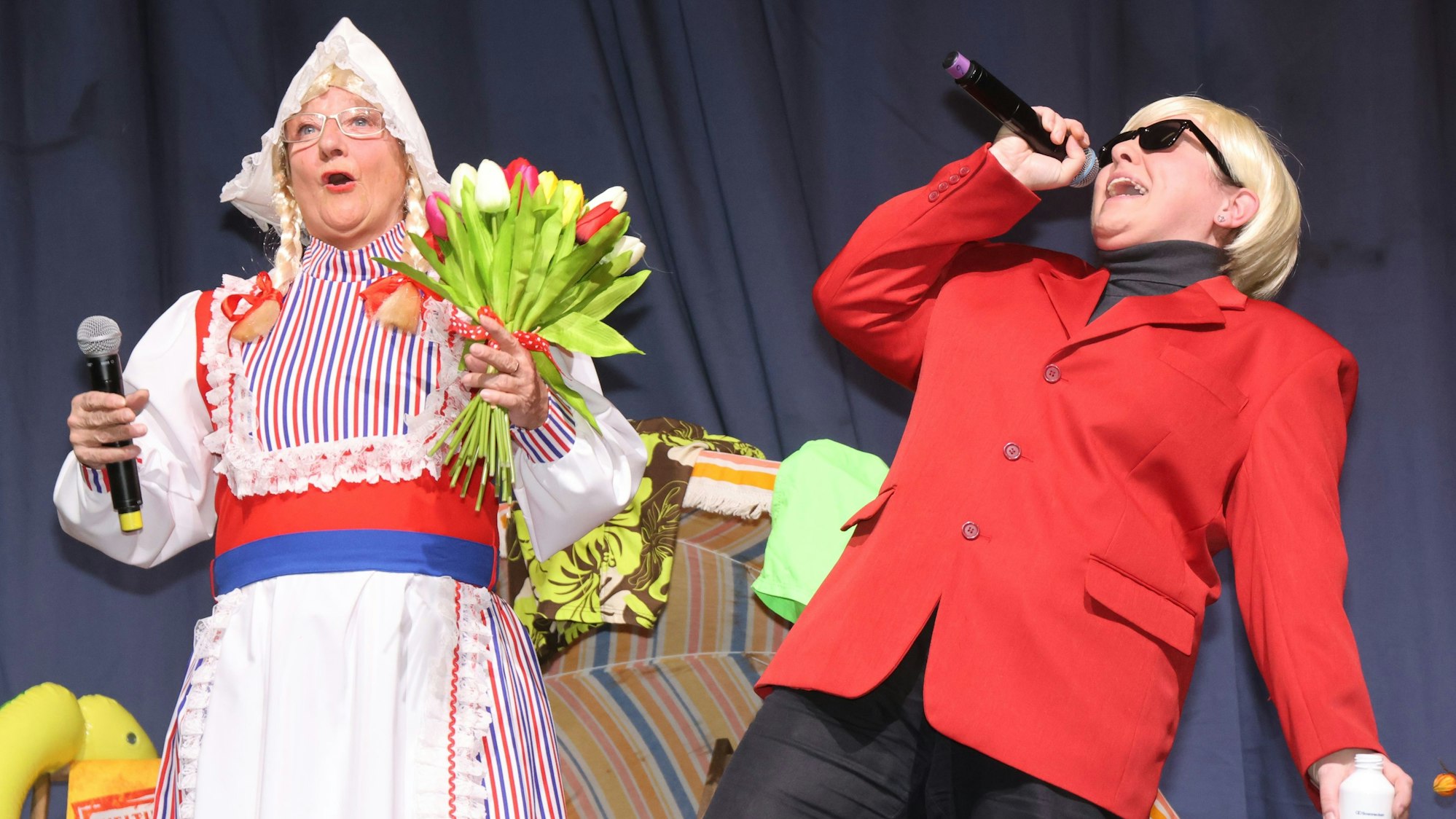 Zwei Frauen stehen auf der Bühne. Eine verkleidet als Frau Antje, mit bunten Tulpen in der Hand, die andere als Heino, der in ein Mikrofon singt.