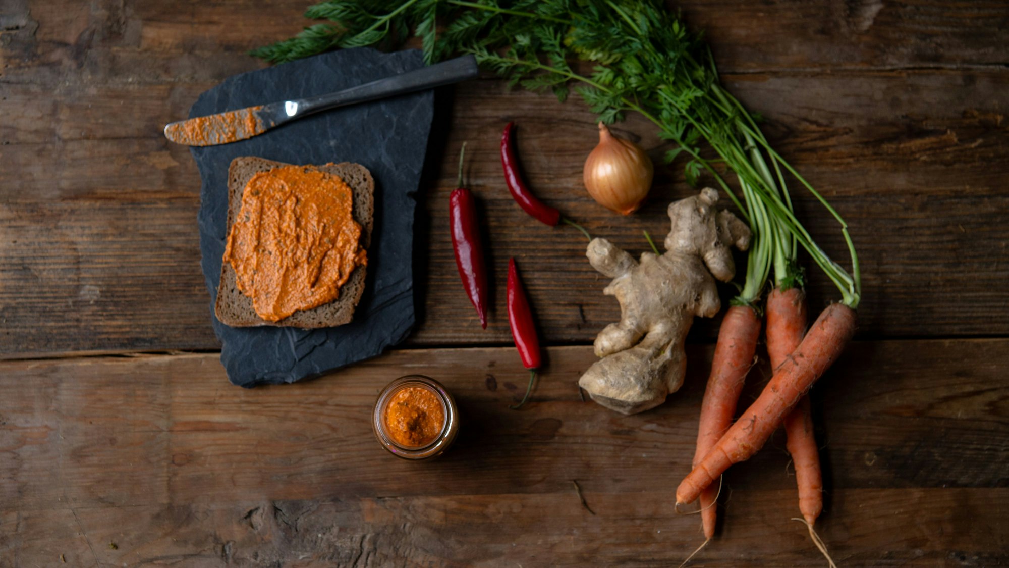 Eine Scheibe Brot, bestrichen mit einem Aufstrich, liegt neben frischem Gemüse in Form von Möhren, Zwiebeln, Chili und Ingwer.