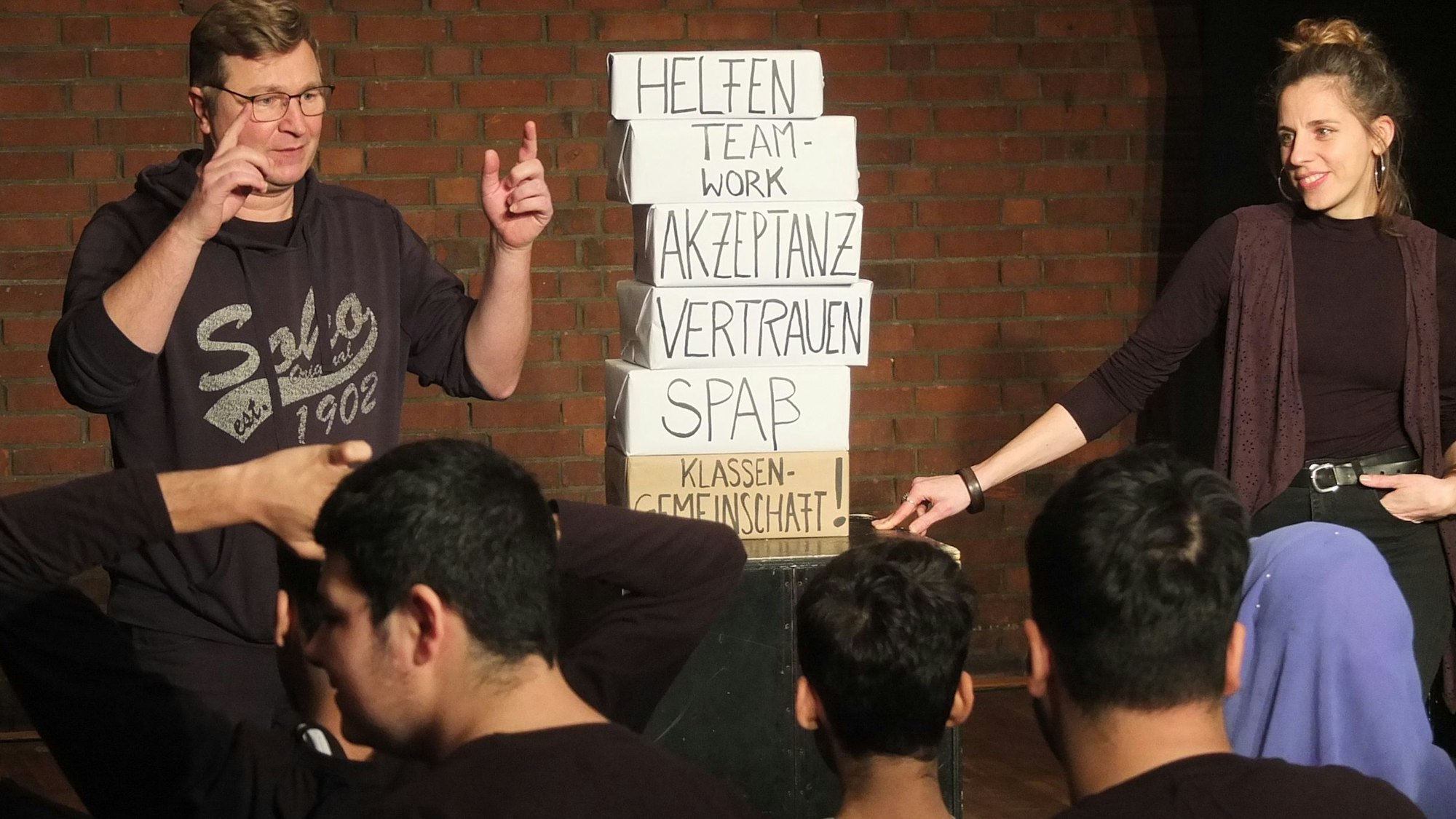 Die Theaterpädagogen Jörg Fabrizius und Anni Auditor stehen vor Schülern und zeigen auf Pappkisten auf denen die wichtigsten Werte eine Klassengemeinschaft stehen: Vetrauen, Spaß und Teamwork zum Beispiel.