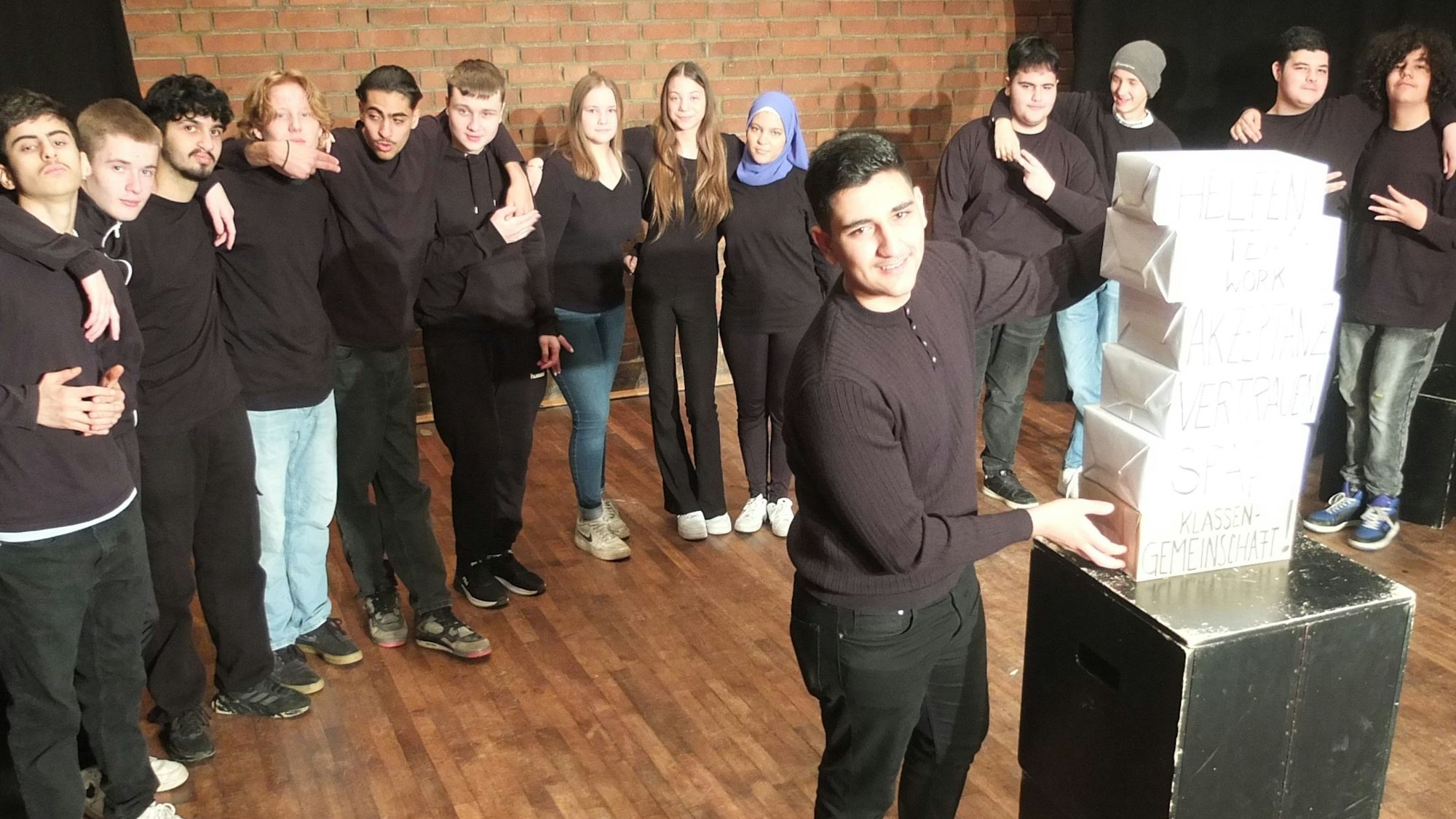 Die jungen Schauspielerinnen und Schauspieler aus der Jahrgangsstufe 9 der Wilhelm-Leyendecker-Schule in Ehrenfeld stehen im Halbkreis auf der Bühne. Ein Schüler steht in der Mitte und stapelt Pappkartons aufeinander, auf denen wichtige Werte einer Klassengemeinschaft stehen.