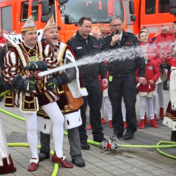 Prinz, Bauer und Jungfrau halten ein Feuerwehrstrahlrohr aus dem Wasser spritzt. Im Hintergrund sind Feuerwehrfahrzeuge zu sehen.