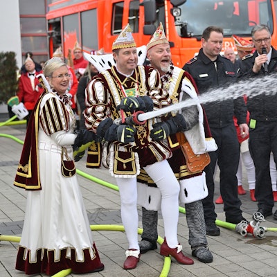 Prinz, Bauer und Jungfrau halten ein Feuerwehrstrahlrohr aus dem Wasser spritzt. Im Hintergrund sind Feuerwehrfahrzeuge zu sehen.