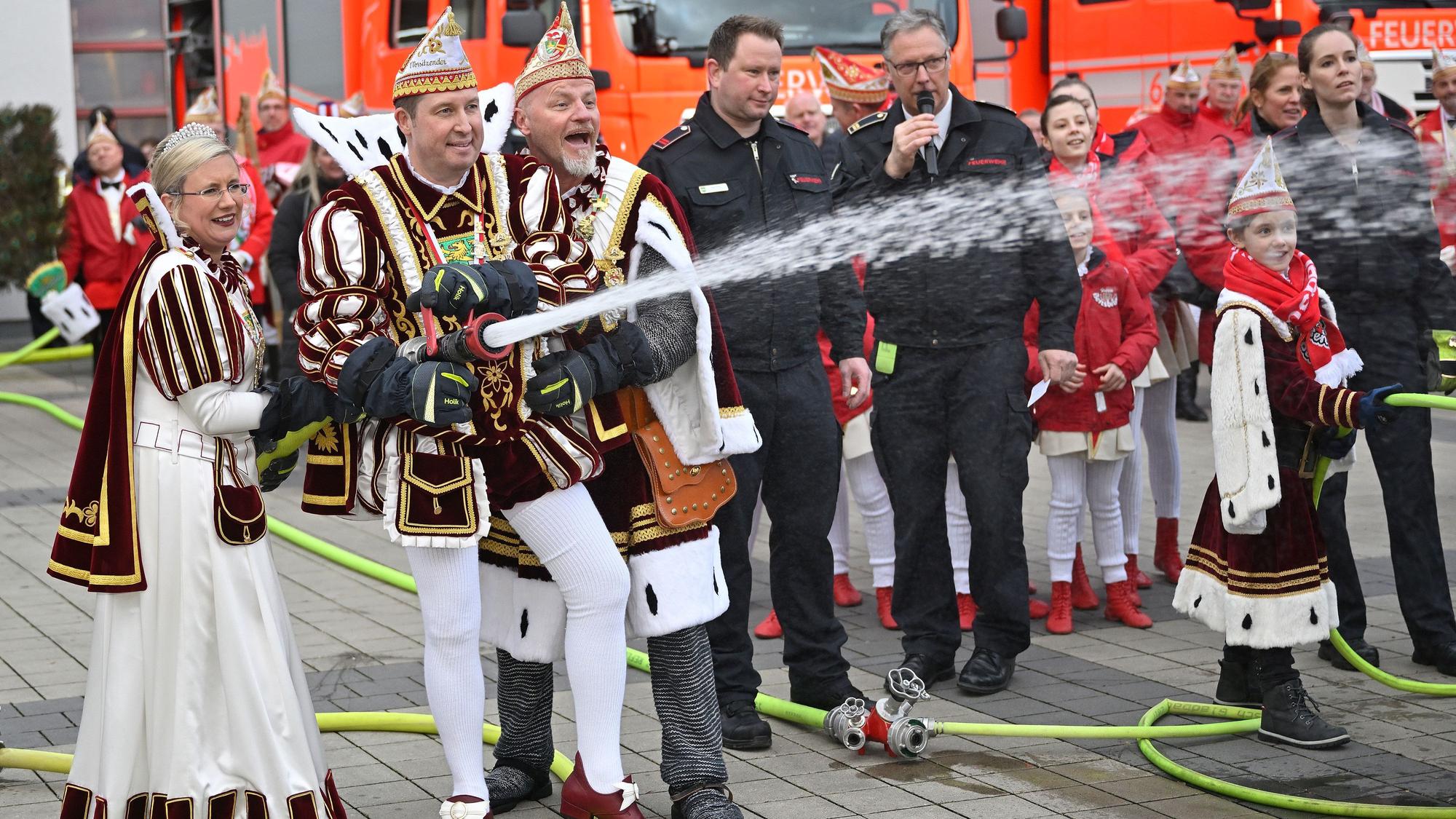 Prinz, Bauer und Jungfrau halten ein Feuerwehrstrahlrohr aus dem Wasser spritzt. Im Hintergrund sind Feuerwehrfahrzeuge zu sehen.