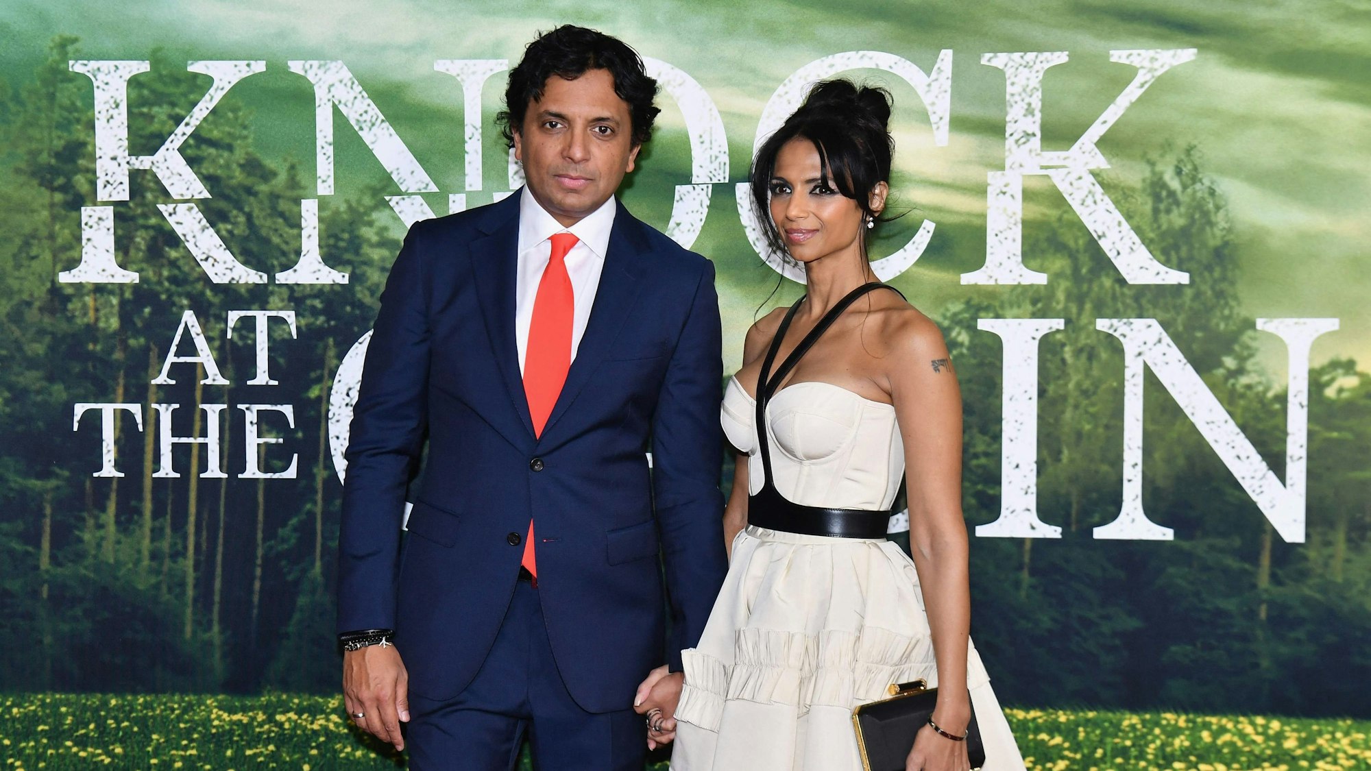 M. Night Shyamalan trägt einen blauen Anzug und eine rote Krawatte, Bhavna Vaswani ein weißes Kleid. Im Hintergrund ist ein Bild zum Film: Eine Blumenwiese, dahinter ein Wald.