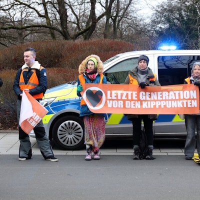 Jugendliche halten ein Transparent mit der Aufschrift „Letzte Generation vor den Kipppunkten“ hoch. Sie stehen am Straßenrand vor einem Polizeiwagen.