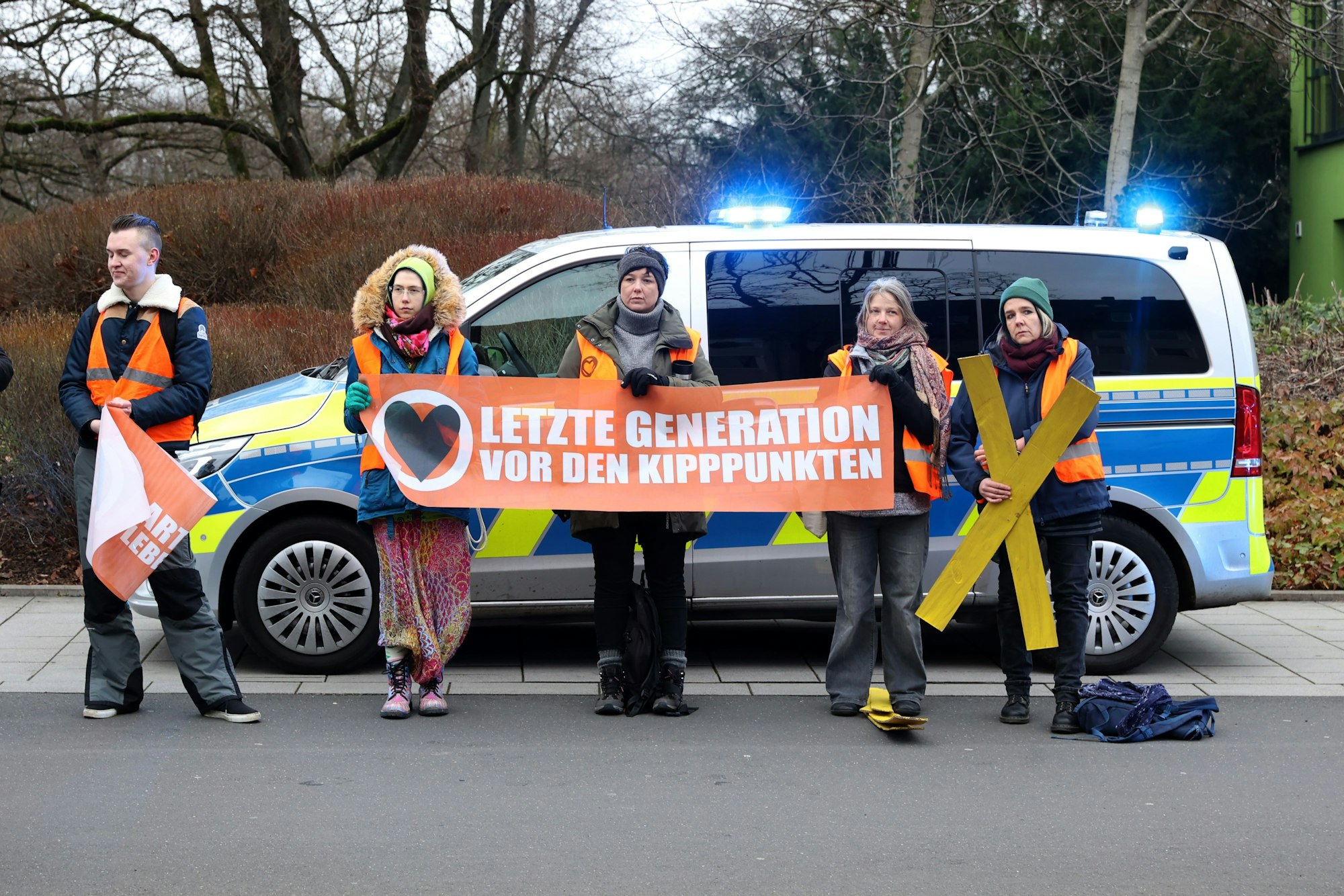 Jugendliche halten ein Transparent mit der Aufschrift „Letzte Generation vor den Kipppunkten“ hoch. Sie stehen am Straßenrand vor einem Polizeiwagen.