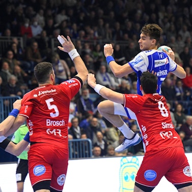 Mehrere Handballspieler zweier Mannschaften sind in einer Spielsituation zu sehen. Einer setzt mit Ball in der Hand zum Sprungwurf an.