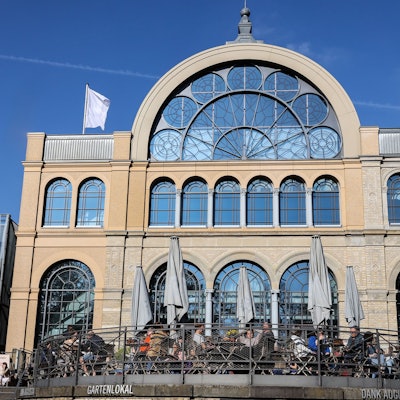 Auf der Terrasse vor dem Kölner Flora-Gebäude im gleichnamigen Botanischen Garten sitzen Menschen an Tischen.