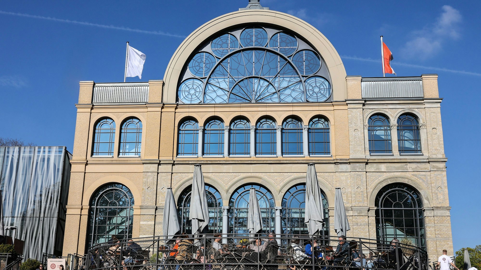 Auf der Terrasse vor dem Kölner Flora-Gebäude im gleichnamigen Botanischen Garten sitzen Menschen an Tischen.