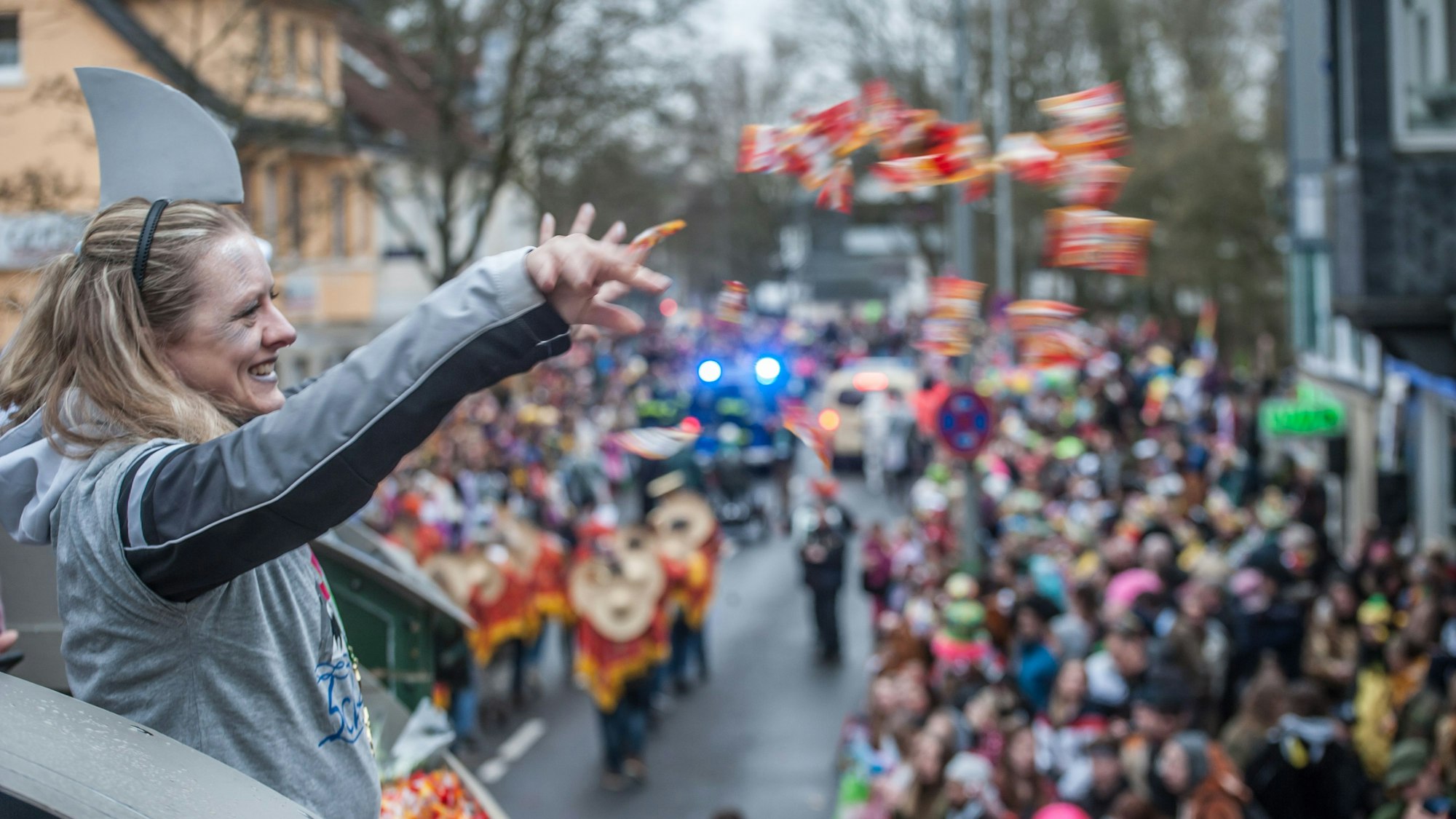 Eine Frau schmeißt eine handvoll Kamelle von einem Wagen in die Zuschauermenge am Wegrand.