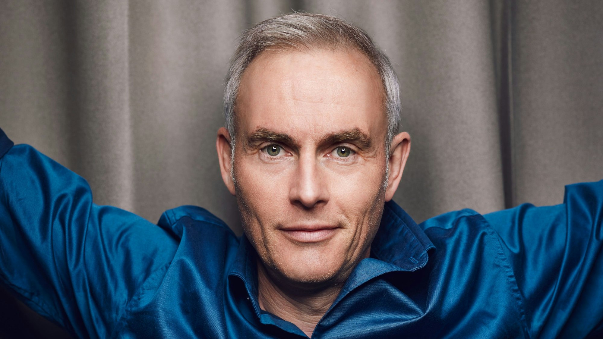 Johann König steht in einem blauen Hemd vor einer grauen Wand.