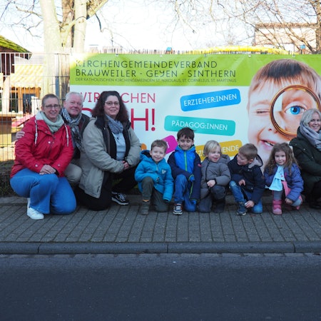 Vor einem Plakat hocken Erwachsene und Kinder auf der Straße. Sie wollen auf Personalnot in Kitas aufmerksam machen.