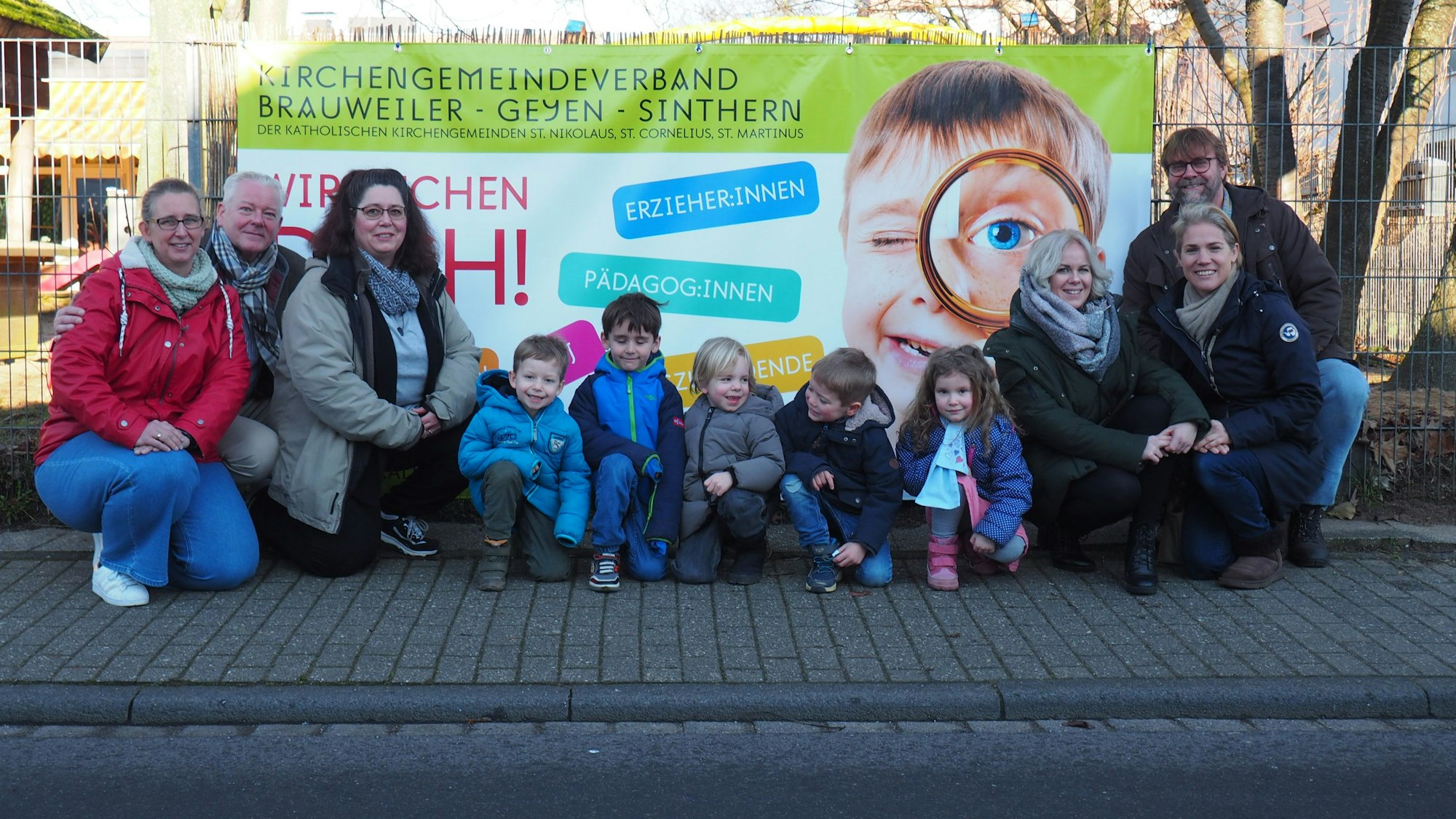 Vor einem Plakat hocken Erwachsene und Kinder auf der Straße. Sie wollen auf Personalnot in Kitas aufmerksam machen.
