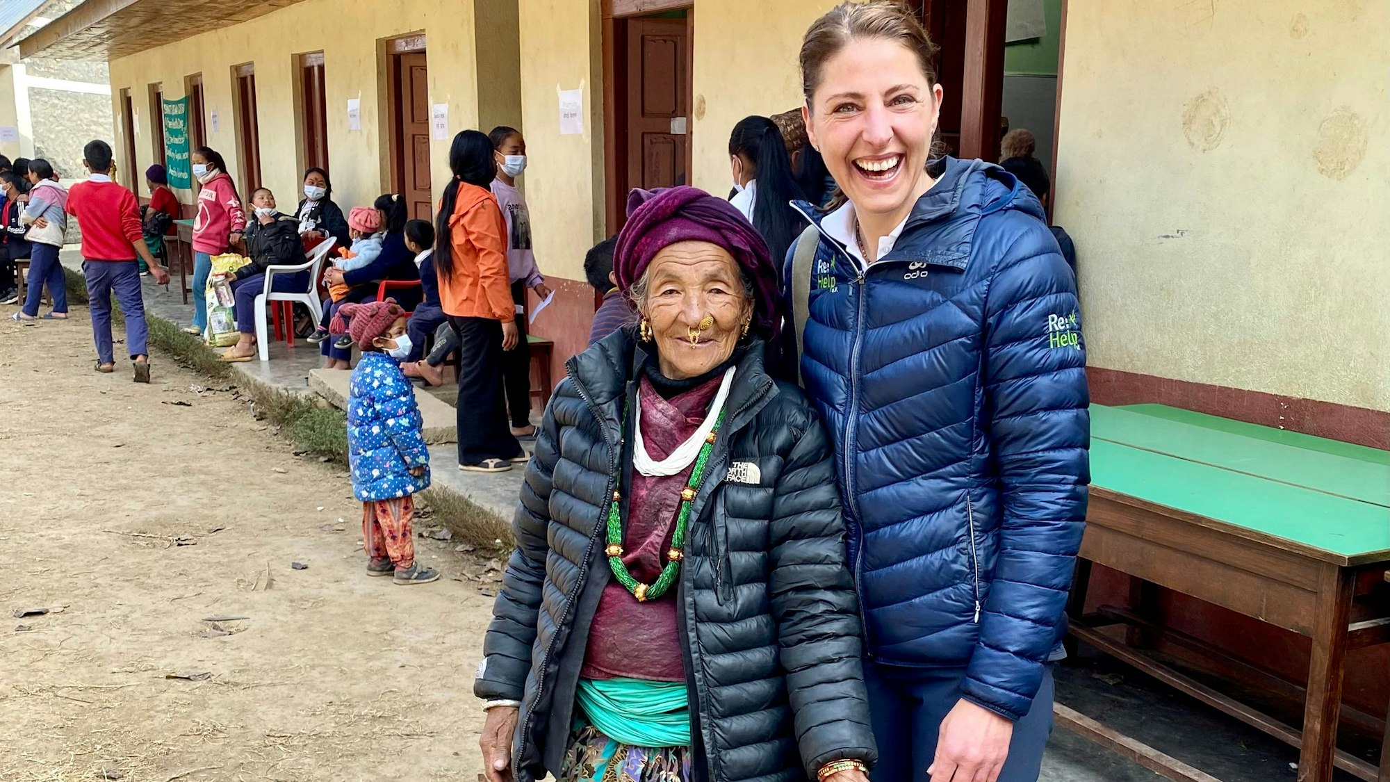 Zwei Frauen, eine jüngere und eine ältere, stehen in einem nepalesischen Ort vor einem gelben Gebäude nebeneinander und lachen in die Kamera