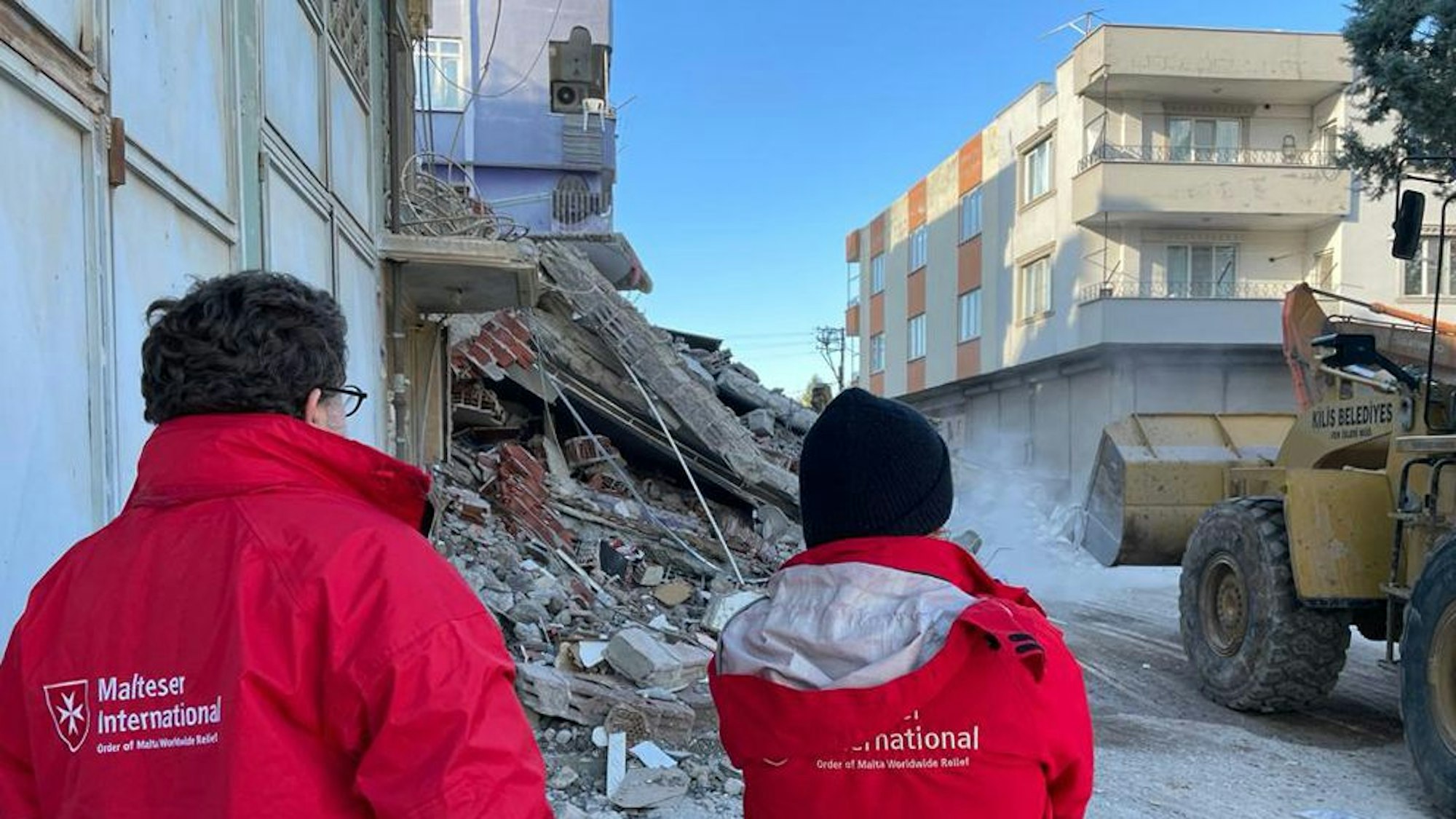 Zwei Personen in roten „Malteser International“-Jacken stehen vor Trümmern, rechts ist ein Bagger zu erkennen. Im Hintergrund steht ein Haus.