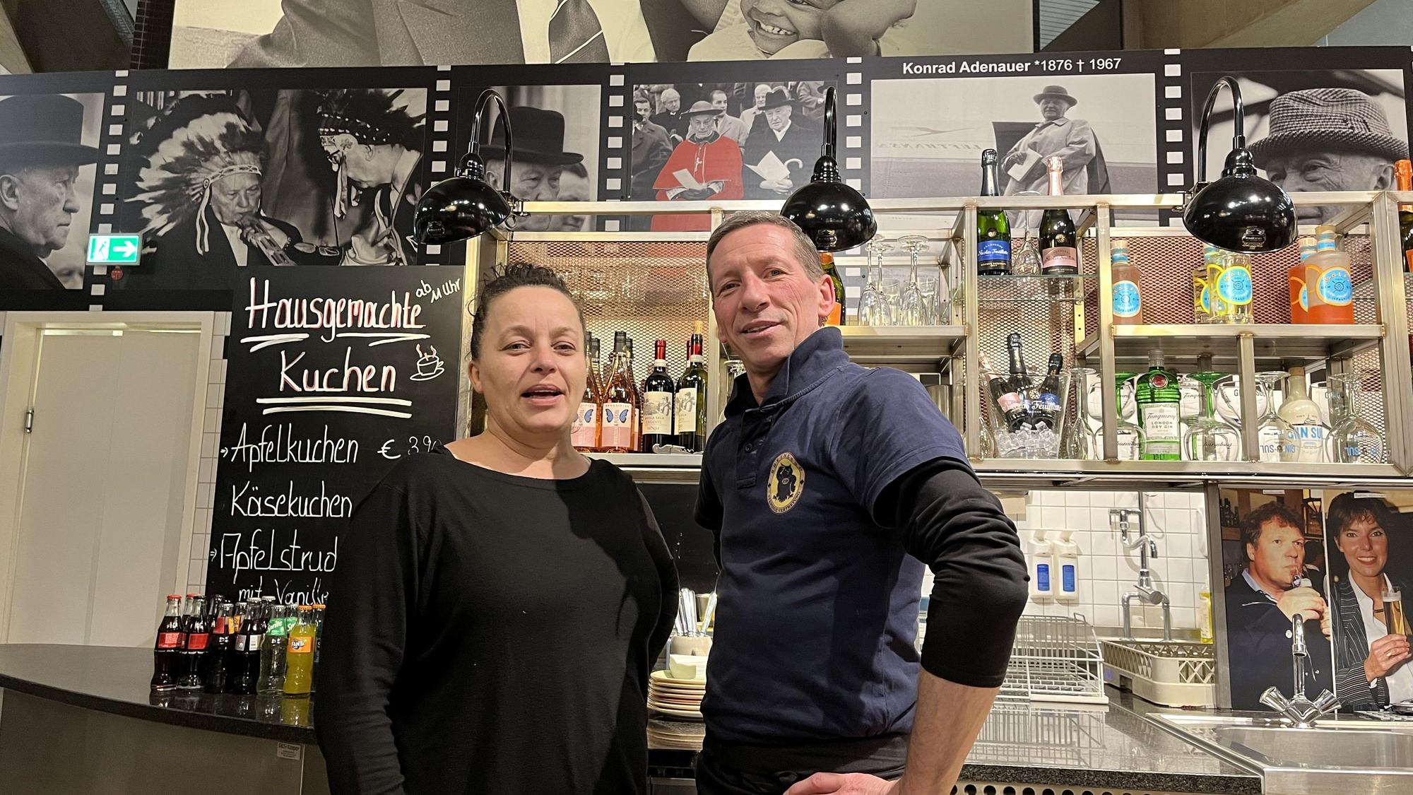 Nopi Kasartziadou und Michael Ebbers arbeiten in dem Lokal „Ständige Vetretung“. Sie stehen an der Theke vor Bildern von Konrad Adenauer und anderen Politikern.