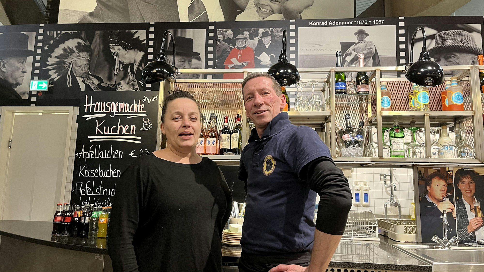 Nopi Kasartziadou und Michael Ebbers arbeiten in dem Lokal „Ständige Vetretung“. Sie stehen an der Theke vor Bildern von Konrad Adenauer und anderen Politikern.