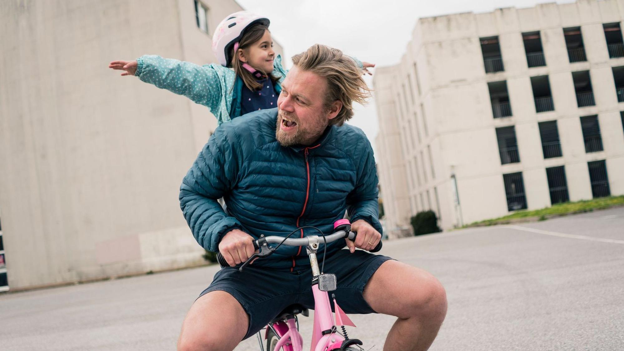 Junger Vater sitzt mit seinem Kind auf einem pinkfarbenen Dreirad (Symbolbild)